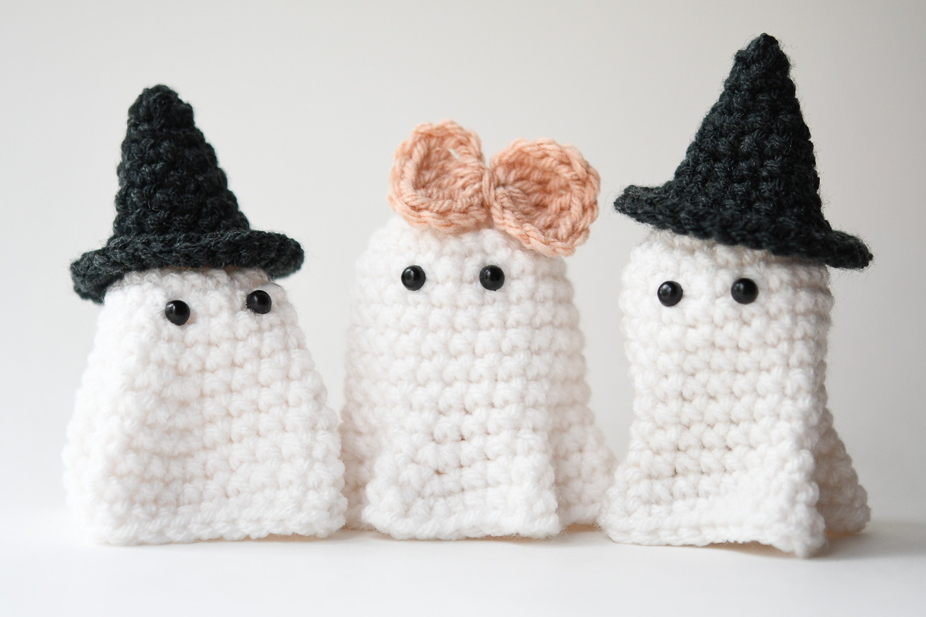 Three crochet ghosts on a white background. Two ghosts are wearing witch hats and one is wearing a pink bow.