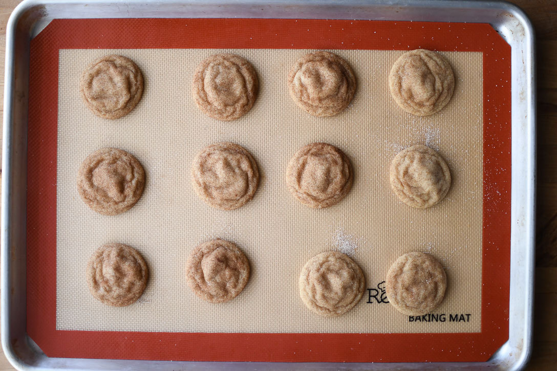 A baking pan lined with a silicone baking mat and twelve freshly baked crinkly snickerdoodles. 