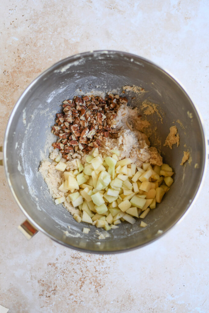 A mixing bowl with apple brownie batter showing apple pieces and pecans being added