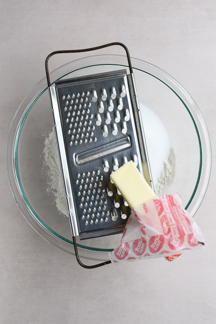 A metal grater with a stick of butter over a mixing bowl